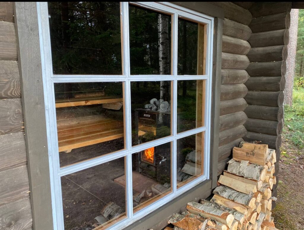Kuva saunan ikkunasta ulkoa sisälle. Ulkona halkoja pinossa, saunan sisällä kiukaassa tuli.